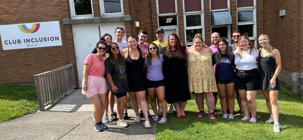 summer camp support team pose together outside of building featuring club inclusion banner